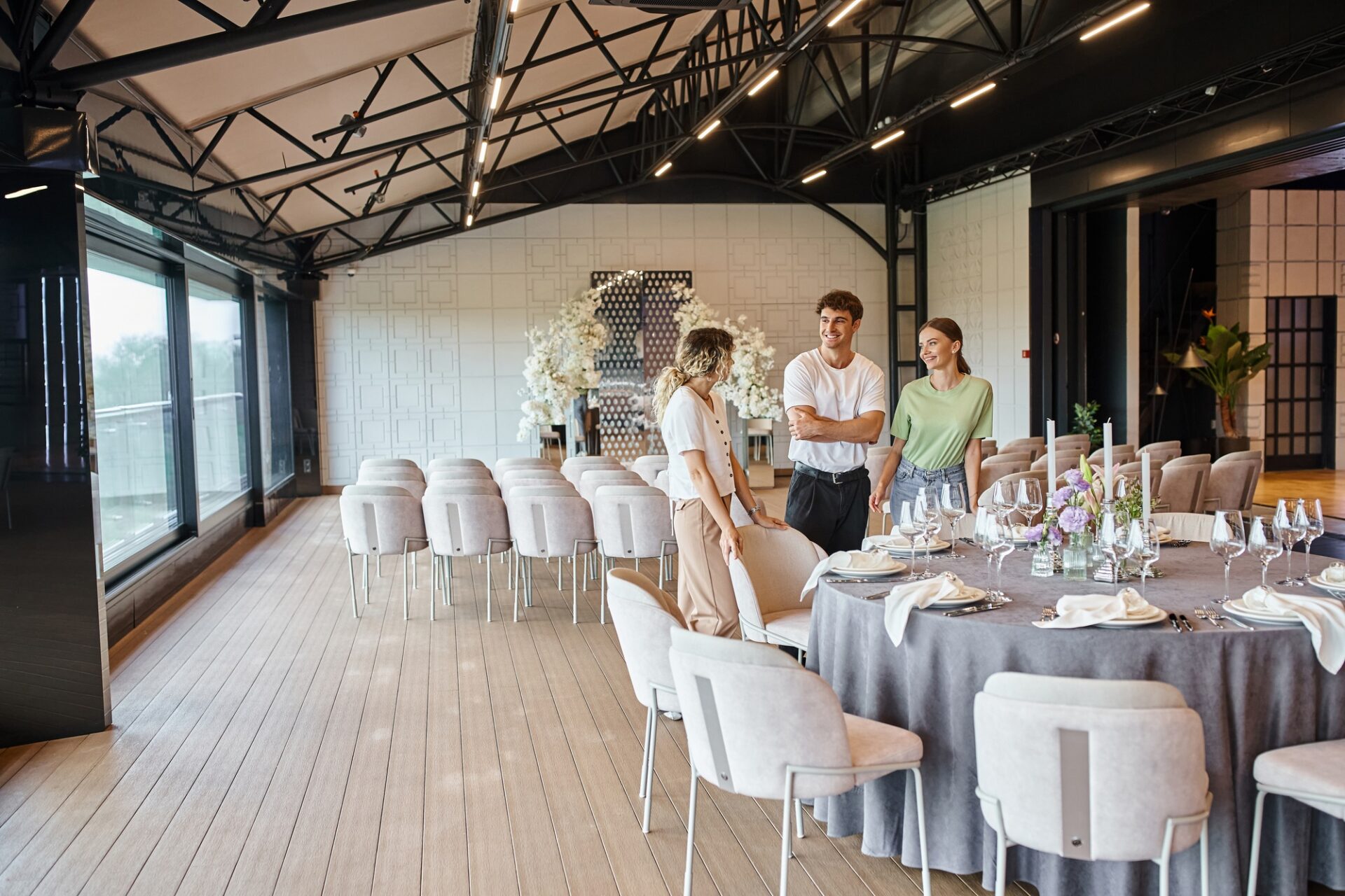 smiling event decorators talking near festive table in spacious decorated event hall, teamwork